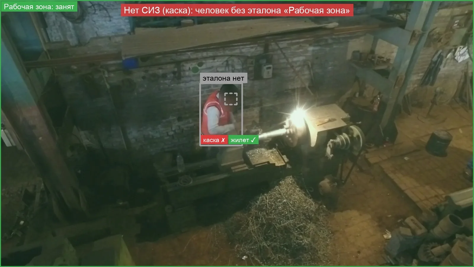 Тёмный механический цех, вид сверху: токарь в сигнальном жилете без каски — нарушение «Нет СИЗ (каска)»; лицо размыто